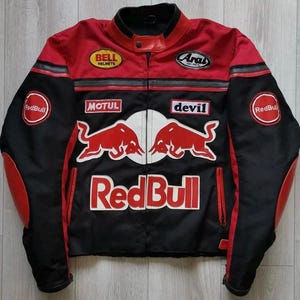 May include: A black and red motorcycle jacket with multiple logos, including "Red Bull," "Bell Helmets," and "Arai." The jacket features a zipper closure, red accents on the shoulders and sleeves, and a stand-up collar.