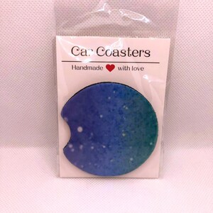 May include: A circular car coaster featuring a blue and teal gradient with white speckles, packaged in a clear plastic sleeve with a white backing card that reads Car Coasters and Handmade with love with a small red heart icon included.