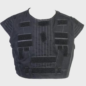 May include: Black vest with multiple rectangular and geometric patches. The vest has short sleeves and is constructed with vertical stitching and a central diamond-shaped patch. The patches are made of a darker material and are attached with velcro.