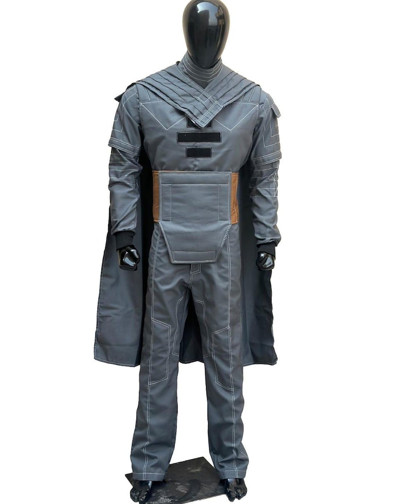 May include: A gray costume with a cape, pants, and a jacket. The jacket has a high collar and a brown belt. White stitching outlines the seams. The costume is displayed on a black mannequin.