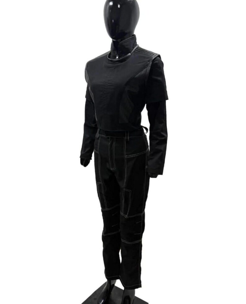 Puede incluir: Conjunto negro de dos piezas sobre un maniqu&iacute;. La parte superior es una camisa de manga corta con cuello alto y una camisa de manga larga debajo. Los pantalones son de cintura alta con detalles de costura blancos. El conjunto es de tela oscura.