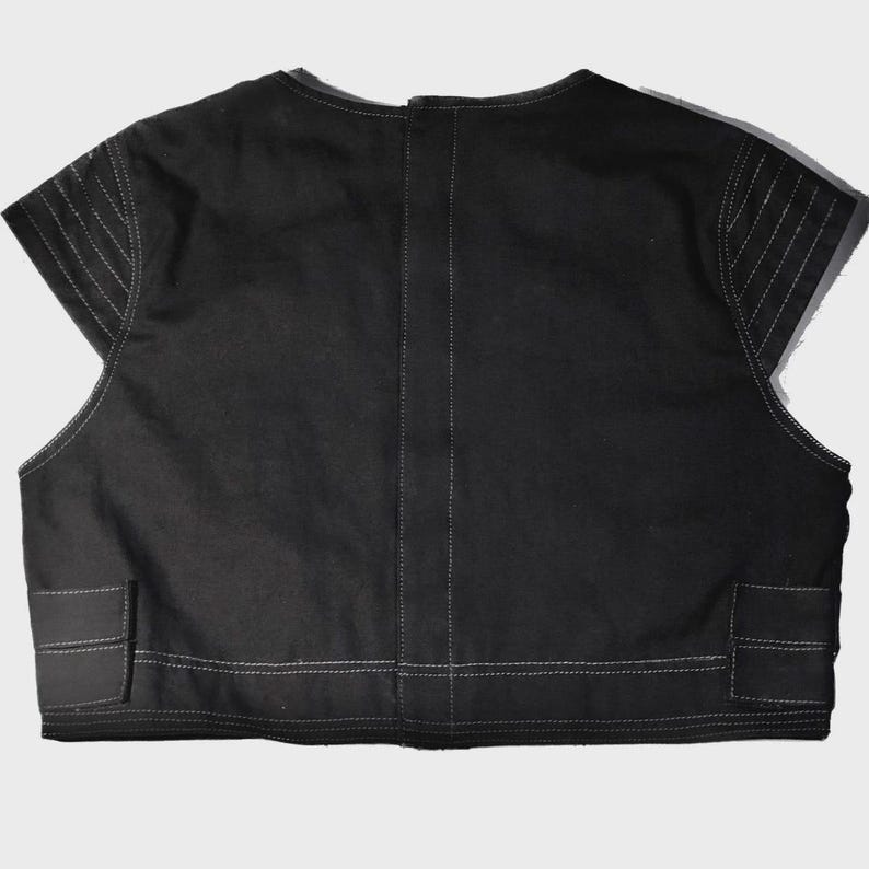 May include: Black vest with a round neckline and short sleeves. The vest features white stitching along the seams and on the shoulders. Two rectangular pockets are visible near the bottom. The vest is made of a sturdy fabric.
