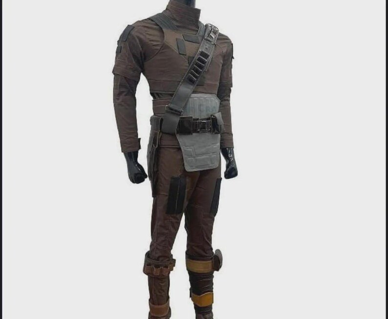 Puede incluir: Un disfraz marr&oacute;n y gris con cuello alto, mangas largas y pantalones a juego. El atuendo incluye una placa pectoral gris, un cintur&oacute;n negro y una bandolera con bolsillos. Guantes negros y botas marrones completan el conjunto.