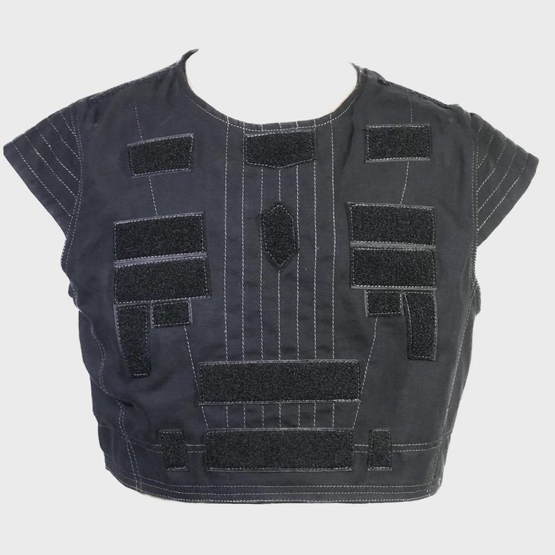 May include: Black vest with multiple rectangular and geometric patches. The vest has short sleeves and is constructed with vertical stitching and a central diamond-shaped patch. The patches are made of a darker material and are attached with velcro.
