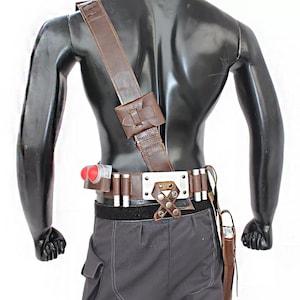 May include: A brown leather utility belt with a matching shoulder strap, pouch, and holster. The belt features silver-colored cylinders and a decorative buckle. A red and silver device is attached to the belt.