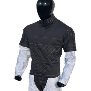 May include: Black short-sleeve shirt with white stitching details and a high collar. The shirt has a geometric design on the front and is layered over a white long-sleeve shirt. The mannequin is wearing white pants.