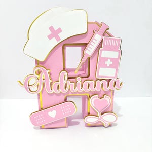 May include: A pink and gold nurse-themed cake topper with the name "Adriana". The design includes a nurse's cap, syringe, pill bottle, bandage, and stethoscope. The topper is designed to look like the letter "A".