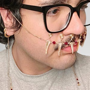 May include: A gold-toned face accessory featuring dangling teeth-like pendants and red beads. The jewelry is connected by delicate chains and wire, with additional decorative elements near the nose and ear. The person is wearing black-framed glasses and a gray shirt.