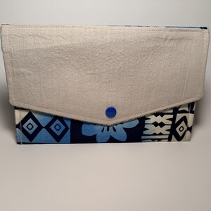 Könnte beinhalten: Eine rechteckige Clutch mit einem cremefarbenen Überschlag, der mit einem blauen Knopf befestigt ist. Der Körper der Clutch weist ein blau-weißes geometrisches Muster auf. Der Überschlag besteht aus einem strukturierten Stoff.