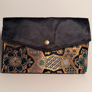 May include: A rectangular clutch bag featuring a solid black fabric flap with a single gold-tone snap button closure. The lower portion of the bag displays a complex, multi-colored woven pattern in shades of teal, gold, and black with geometric motifs.