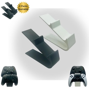 May include: Black and white game controller stands. The stands are angled with a curved base and a raised section to hold the controller. The image shows two stands, one black and one white, with the text "2-Packs Available".