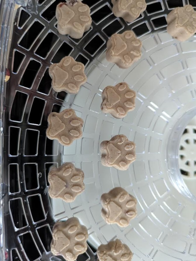 May include: Close-up of several light brown, paw-shaped treats arranged on a clear, plastic dehydrator tray. The treats have a detailed paw print design. The tray has a grid pattern and is slightly curved.