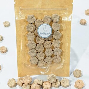 May include: A clear plastic bag filled with paw-shaped treats, with a brown paper backing. The treats are light brown and scattered around the bag. A circular sticker on the bag reads "Hop Harvest" with a bunny logo.