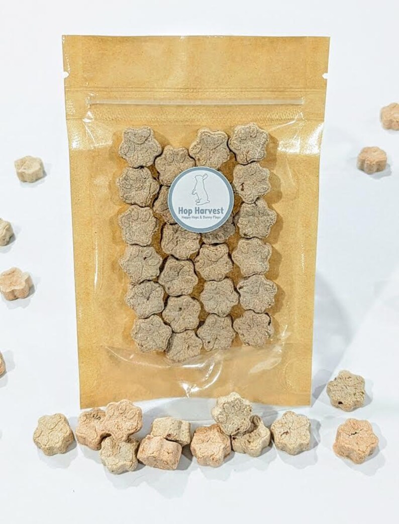 May include: A clear plastic bag filled with paw-shaped treats, with a brown paper backing. The treats are light brown and scattered around the bag. A circular sticker on the bag reads "Hop Harvest" with a bunny logo.