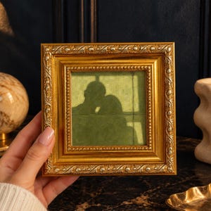 May include: A square picture frame with a metallic gold finish and ornate, embossed leaf and bead detailing. The frame contains a painting of a dark silhouette of two people against a muted olive green background with a window pane grid.