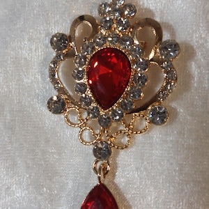 May include: A gold-tone metal brooch featuring a central pear-shaped red gemstone surrounded by clear rhinestones in a decorative filigree frame, with a matching pear-shaped red gemstone dangling from the bottom of the piece.