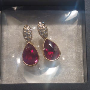 May include: A pair of gold-tone drop earrings featuring a pear-shaped deep red gemstone surrounded by a thin metal border. Above each red stone is a marquise-shaped accent piece encrusted with small, clear, sparkling white stones.