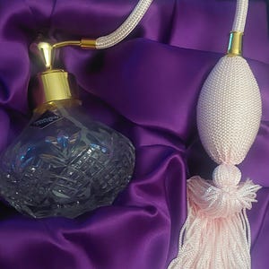 May include: A clear cut-glass perfume bottle with a gold-tone metal collar and a light pink fabric-covered atomizer bulb. The bulb features a matching light pink tassel and is connected to the bottle by a flexible, fabric-covered hose.