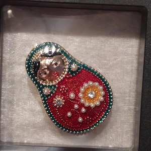May include: A Russian nesting doll brooch featuring a red body with a green border, decorated with small white pearls and a central yellow flower motif with a clear crystal centre. The doll has a metallic face with black hair and red lips.