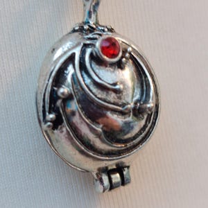 May include: A silver-toned metal locket pendant with a rounded, layered design featuring curved lines and small spherical accents. A single, small, circular red gemstone is embedded near the top of the locket, which includes a hinged closure.