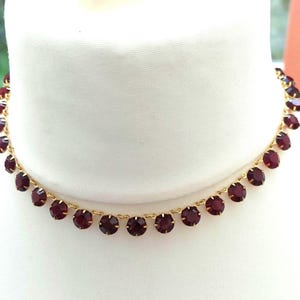 May include: A gold-toned choker necklace featuring a continuous row of round, deep red faceted stones set in individual metal prongs. The necklace is displayed on a white form, showcasing the uniform arrangement of the dark red gems and the metallic gold chain links connecting each setting in a circular design.