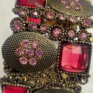 May include: A vintage-style bracelet with a bronze finish. The bracelet features large, faceted, ruby-red rectangular and square stones, interspersed with smaller pink gemstones. The design includes ornate, textured metalwork and circular accents, creating a bold, statement piece.