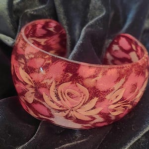 May include: A wide, translucent cuff bracelet featuring a deep red and pink mottled pattern. The surface is decorated with an embossed, light-coloured floral design consisting of a central rose surrounded by leaves that wrap around the band.