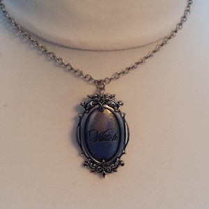 May include: A silver-toned oval pendant necklace featuring an ornate, filigree-style frame surrounding a dark, smooth center. The word Witch is inscribed in a cursive script across the middle of the pendant, which hangs from a simple silver chain.