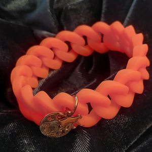 Könnte beinhalten: Ein leuchtend neon-oranges Gliederkettenarmband mit matter Oberfläche. Ein kleiner, runder, goldfarbener Metallanhänger hängt an einem der Glieder und zeigt eine geprägte Grafik einer stilisierten Tierfigur sowie einige undeutliche Buchstaben.