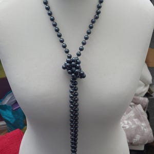 May include: A long necklace featuring dark grey, metallic-finish spherical beads strung on a dark cord. The necklace is tied into a central knot, creating a lariat-style design that hangs down the centre of the chest with two beaded strands.