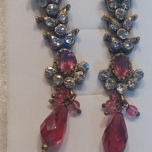 May include: A pair of long drop earrings featuring an antique gold-tone metal finish. Each earring has a vertical row of clear round rhinestones, a central oval pink stone surrounded by smaller clear rhinestones, and a dangling red teardrop crystal.