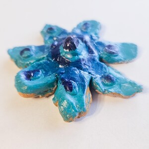 May include: A handcrafted, star-shaped ornament with a textured surface. The ornament features a blend of blue and turquoise hues, with darker blue accents. The edges have a natural, unpainted appearance.