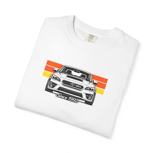 May include: White t-shirt with a car illustration in black and white, text reading "WRX" and "Since 1992". The design includes orange and yellow stripes. The shirt is folded.