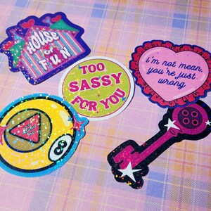 May include: A collection of colorful stickers with glitter accents. Designs include a house with "HOUSE OF FUN" text, a round sticker with "TOO SASSY FOR YOU", a heart with "I'm not mean, you're just wrong", an eight ball, and a key.