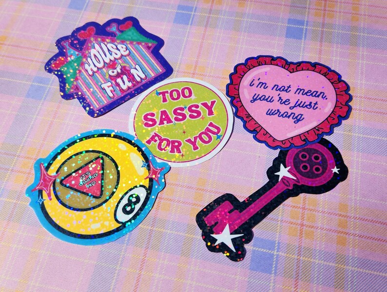 May include: A collection of colorful stickers with glitter accents. Designs include a house with "HOUSE OF FUN" text, a round sticker with "TOO SASSY FOR YOU", a heart with "I'm not mean, you're just wrong", an eight ball, and a key.