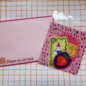 May include: Pink envelope with a scalloped edge and the text "Please do not bend". A pink card with a strawberry and a bear graphic is in a clear plastic bag. The card has the text "Life is Happy OS".