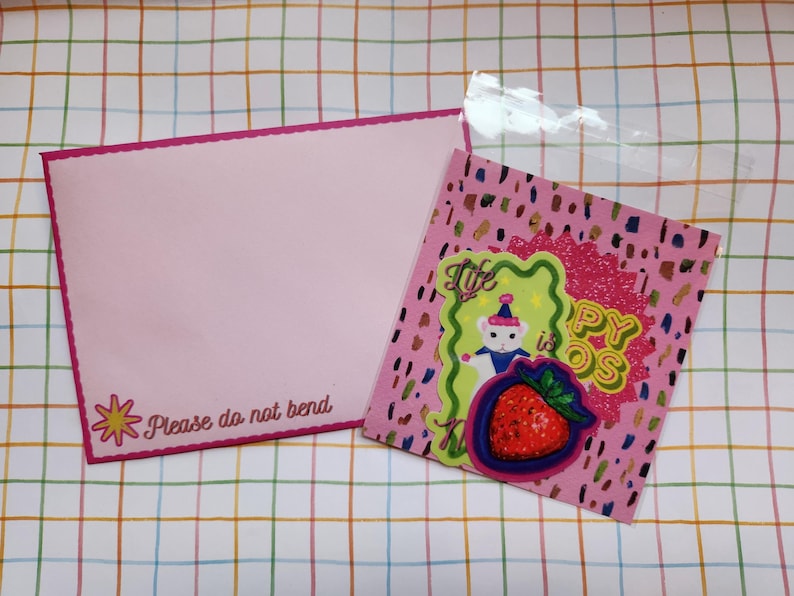 May include: Pink envelope with a scalloped edge and the text "Please do not bend". A pink card with a strawberry and a bear graphic is in a clear plastic bag. The card has the text "Life is Happy OS".