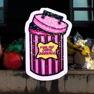 May include: A pink and black sticker in the shape of a trash can with the text "FOR MY JUNK JOURNAL" on a yellow starburst. The trash can has vertical stripes and glitter accents. The background is a blurry outdoor scene.