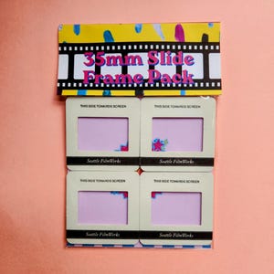 Könnte beinhalten: Eine Packung mit 35-mm-Dias. Die Verpackung ist gelb mit bunten Pinselstrichen und dem Text "35mm Slide Frame Pack". Vier Diarahmen sind sichtbar, jeweils mit dem Text "This Side Towards Screen" und "Seattle FilmWorks".