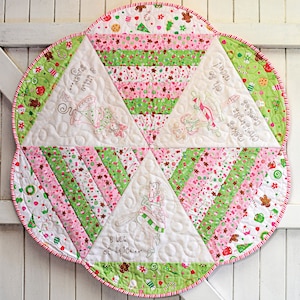 May include: A white quilted table runner with a scalloped edge. The runner is decorated with a green and pink Christmas-themed fabric with gingerbread men, candy canes, and other holiday motifs. The runner features four embroidered images of fairies with text that reads "a lot of spice...", "Christmas Cookies oh so nice!" and "a little bit of magic".