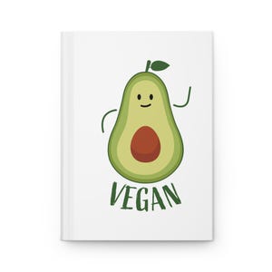 May include: White notebook featuring a cartoon avocado illustration. The avocado is green with a brown pit, a smiling face, and the word "VEGAN" in green below. The notebook has a clean, minimalist design.