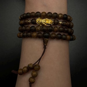 May include: A multi-strand bracelet featuring dark brown wooden beads and a gold-colored decorative element. The bracelet is strung on a dark brown cord and is displayed on a person's arm. The beads are approximately 0.8 cm in diameter.