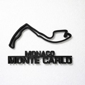 May include: A black, three-dimensional wall art piece featuring the outline of the Monaco Grand Prix circuit. Below the track layout, the text MONACO and MONTE CARLO are displayed in bold, raised, black capital letters with a textured finish.