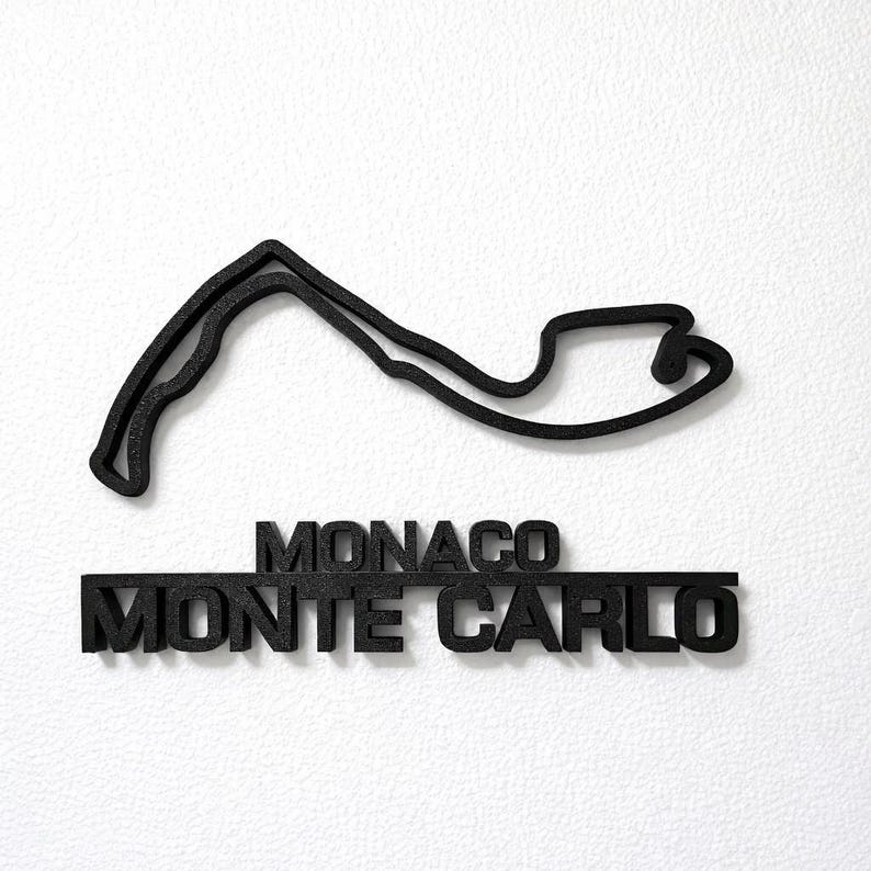 May include: A black, three-dimensional wall art piece featuring the outline of the Monaco Grand Prix circuit. Below the track layout, the text MONACO and MONTE CARLO are displayed in bold, raised, black capital letters with a textured finish.