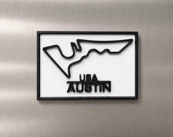 Può includere: Una targa da parete rettangolare con un contorno nero in rilievo del circuito di gara Circuit of the Americas su sfondo bianco. Sotto il disegno del tracciato, il testo USA AUSTIN è visualizzato in lettere maiuscole nere, in grassetto e in rilievo.