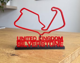 Può includere: Una scultura rossa stampata in 3D del tracciato del circuito di Silverstone, montata su una base rettangolare nera. Il testo UNITED KINGDOM SILVERSTONE è impresso in lettere maiuscole rosse sulla parte anteriore della base sotto il circuito.