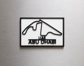 Può includere: Una targa da parete rettangolare con un contorno nero in 3D del circuito di Formula 1 di UAE Abu Dhabi su sfondo bianco. Il testo UAE ABU DHABI è visualizzato in lettere nere in 3D sotto il design del tracciato del circuito.