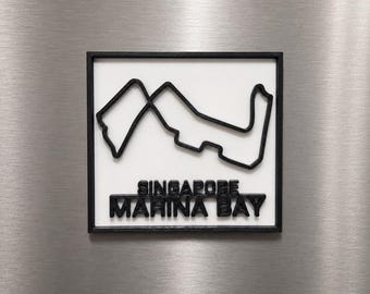 Può includere: Una targa da parete quadrata con un contorno nero in rilievo del circuito cittadino di Marina Bay a Singapore su sfondo bianco. Il testo SINGAPORE MARINA BAY è impresso in lettere nere sotto il tracciato, il tutto racchiuso in una cornice nera.