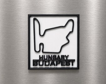 Può includere: Una targa da parete quadrata con un contorno nero in rilievo del circuito di gara Hungaroring su sfondo bianco. Sotto il disegno del tracciato, le parole HUNGARY e BUDAPEST sono impresse in lettere maiuscole nere entro una cornice nera.
