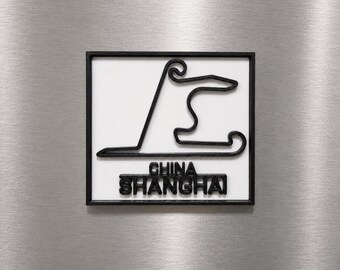 Può includere: Una targa da parete quadrata con un contorno nero in rilievo del tracciato del circuito internazionale di Shanghai su sfondo bianco. Il testo CHINA SHANGHAI è impresso in lettere nere sotto la mappa del circuito, il tutto incorniciato da un bordo nero.